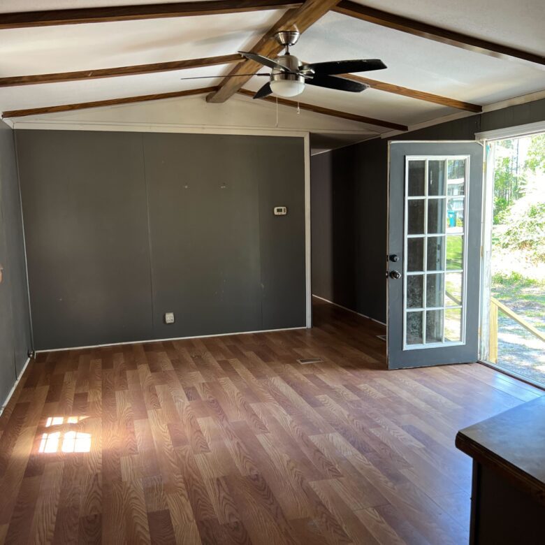 A modern living room featuring dark gray walls, hardwood flooring, and wooden ceiling beams. The room includes large windows with curtains and French doors that open to a scenic outdoor view. A ceiling fan with a light fixture is centrally mounted on the ceiling.