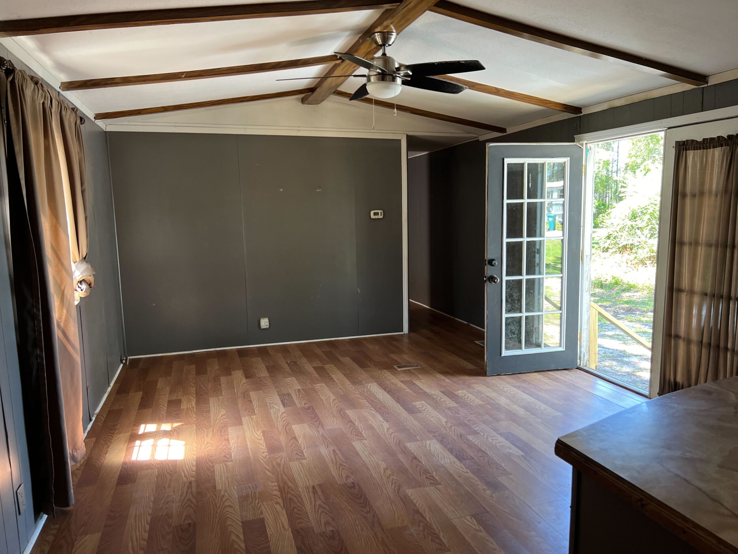 A modern living room featuring dark gray walls, hardwood flooring, and wooden ceiling beams. The room includes large windows with curtains and French doors that open to a scenic outdoor view. A ceiling fan with a light fixture is centrally mounted on the ceiling.