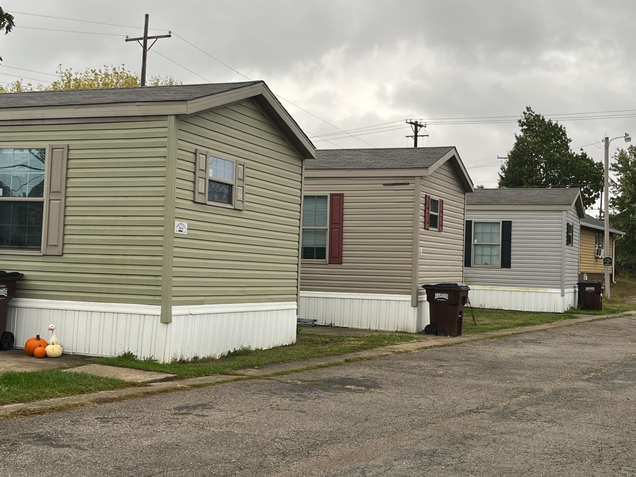 Street with mobile homes on one side, featuring well-maintained units with front porches and small gardens, cloudy sky overhead