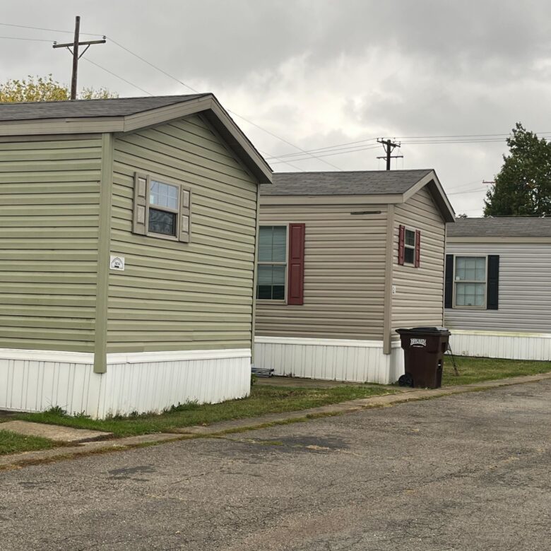 Street with mobile homes on one side, featuring well-maintained units with front porches and small gardens, cloudy sky overhead