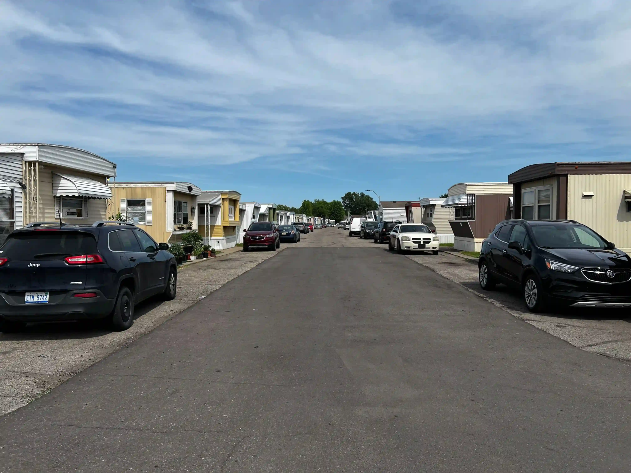 Main street view of Roseville Mobile Home Park with mobile homes on both sides, cars parked along the curb, and a clear sky overhead.