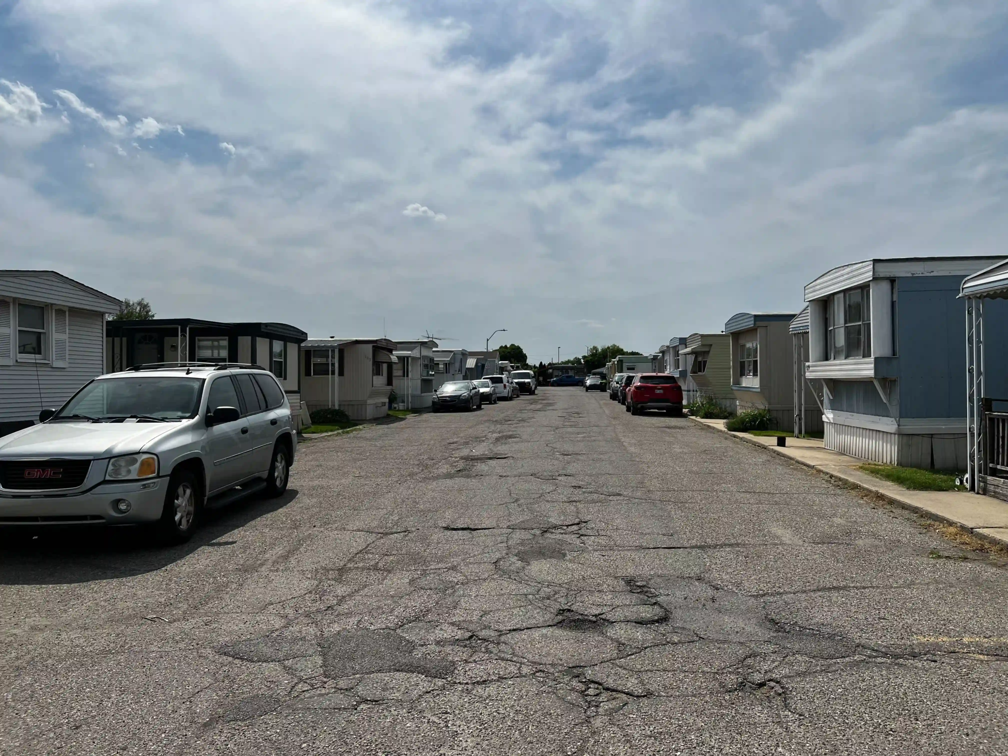 Street view in Roseville Mobile Home Park with parked vehicles, mobile homes on either side, and a partly cloudy sky overhead.